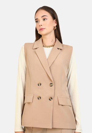 Beige double-breasted vest with notched lapels, four pockets, and smooth fabric. Worn over a light-colored long-sleeve top.