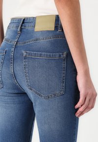 Sötétkék farmernadrág, melynek testhez simuló fazonja van, hátul lapos zsebbel és világoszöld bőrcimkével a deréknál.