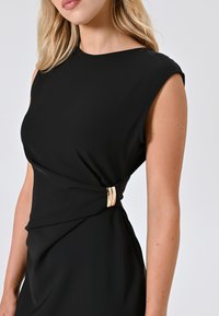 Robe noire au design ajusté, à manches courtes, avec un détail torsadé à la taille et un fermoir en accent doré. Texture de tissu lisse.