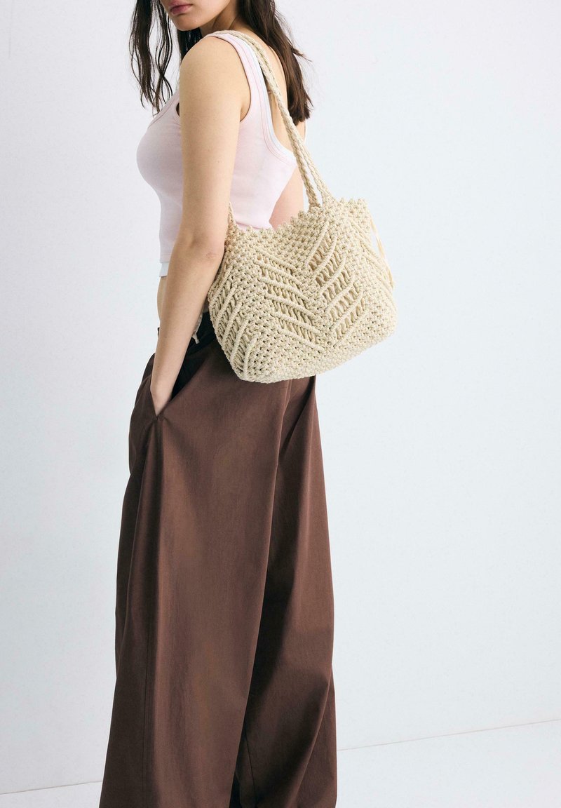 Femme portant un haut blanc sans manches et un pantalon large marron, avec un sac en crochet crème texturé sur l'épaule droite.