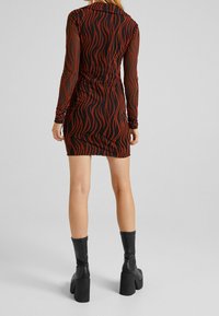 Kobieta w dopasowanej, długiej sukience mini w czarne i rdzawobrązowe faliste paski, w połączeniu z czarnymi trzewikami na platformie, stojąca tyłem.