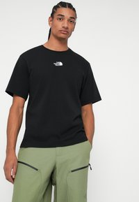 Tricou negru din bumbac cu guler rotund, mâneci scurte și un logo alb pe piept. Asociat cu pantaloni verzi prevăzuți cu buzunare cu fermoar.
