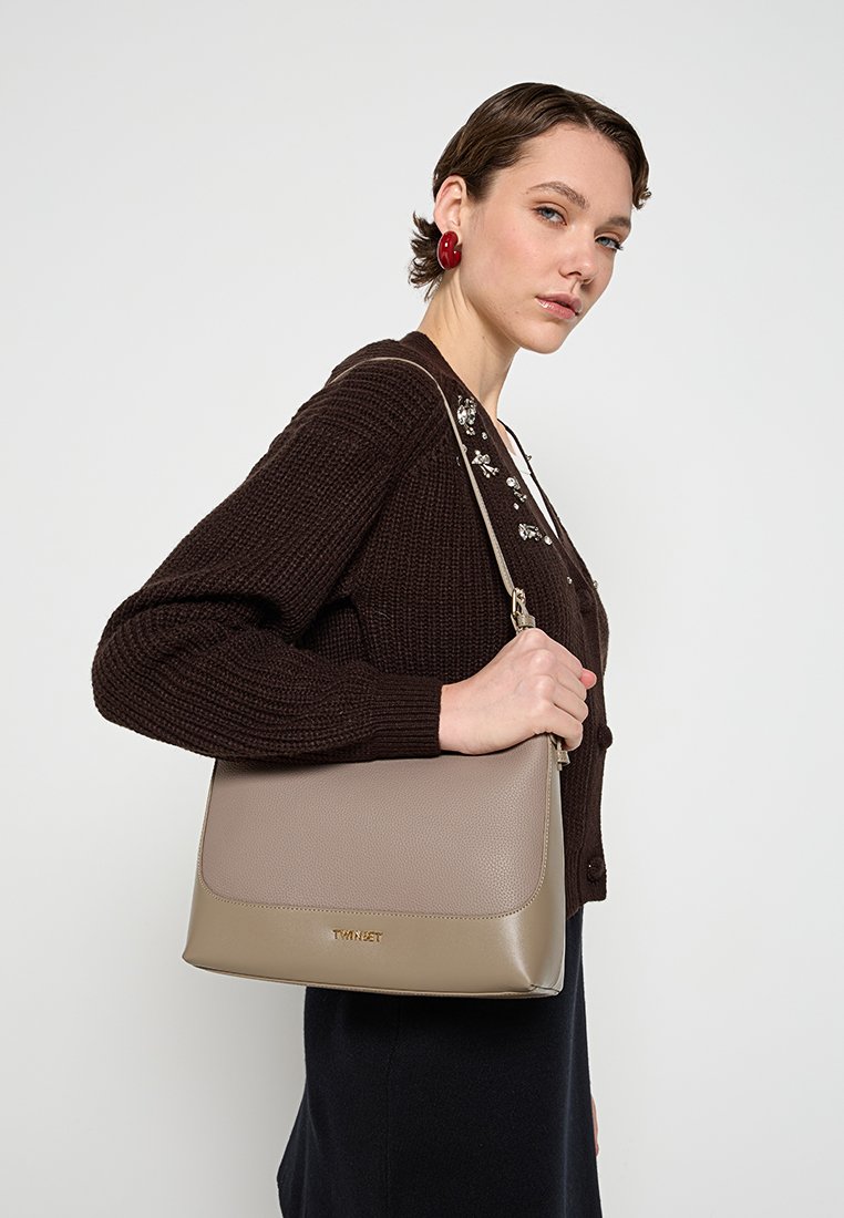 Beige läder axelväska med en strukturerad form, med en slät yta och subtil logotypdetalj på framsidan. Justerbar rem ingår.