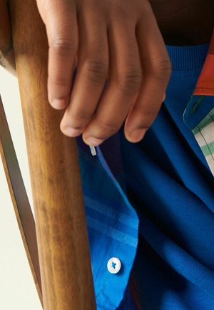 Main tenant une canne en bois, portant une chemise à carreaux bleue avec un bouton blanc, suggérant un soutien ou une aide à la mobilité.