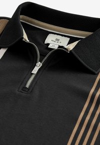 Polo noir zippé avec un col en maille, des accents tan, et trois bandes tan sur le côté gauche. Étiquette de la marque à l'intérieur du col.