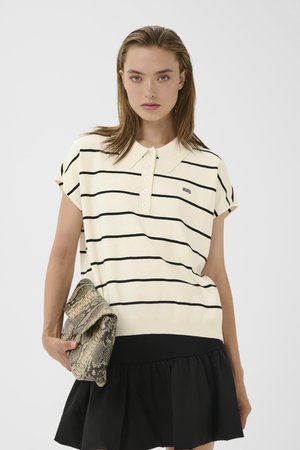Nuori nainen kermanvärisessä ja mustavalkoisessa raitaisessa poolopaidassa ja mustassa hameessa, kädessään käärmeen kuviollinen clutch, valkoista taustaa vasten.