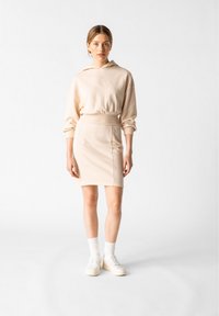 Beige Kapuzenpullover kombiniert mit einem figurbetonten beigen Rock. Das Outfit zeichnet sich durch weiche Texturen und ein minimalistisches Design aus. Weiße Sneakers runden den Look ab.