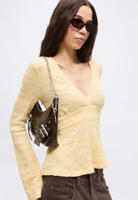 Ung kvinne iført gul, strukturert V-halsgenser, mørke solbriller, brune bukser, og bærer en liten svart skinnveske med sølvkjede over skulderen.