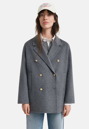 Grau melierte Woll-Doppelskinnjacke mit goldenen Knöpfen, großen Revers, seitlichen Taschen und lockerem Schnitt. Getragen über einem Hemd mit Kragen und Jeans.