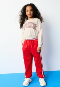 Sweatshirt in hellbeige mit dem Wort "HAPPINESS" in fettem, kastanienbraunem Text, kombiniert mit roten Jogginghosen mit weißen Seitenstreifen und Druckknopfdetails.