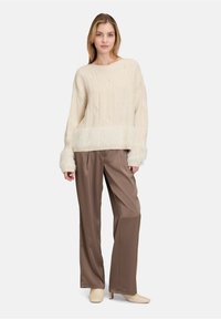 Cremfarbener gestrickter Pullover mit strukturierten Mustern und einem Besatz aus Kunstfell am Saum und an den Bündchen, kombiniert mit weit geschnittenen braunen Satin-Hosen.