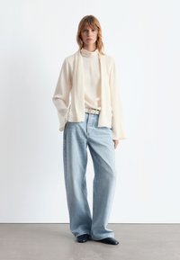 Beige Langarmoberteil mit Stehkragen, kombiniert mit hellblauen weiten Jeans und dunkelblauen spitzen Schuhen. Schlicher weißer Hintergrund.