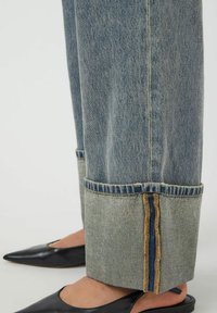 Jeans de mezclilla azul claro enrollados, acompañados de unos zapatos de niña negros con punta afilada y tira en el talón, mostrando un primer plano de la zona de la parte inferior de la pierna.