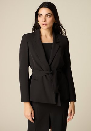 Mujer con cabello largo y oscuro, vestida con un blazer negro con cinturón y falda a juego, posando frente a un fondo liso beige.