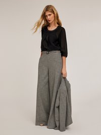 Blusa nera trasparente con maniche 3/4, abbinata a pantaloni a gamba larga grigi a quadri con vita alta. Il modello tiene in mano un blazer grigio coordinato.