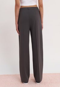 Wide-leg trousers in dark gray fabric, featuring a smooth texture, high waistband, and back pockets, resting on a textured pink surface.