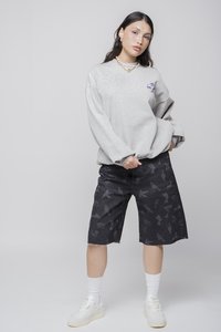 Sudadera gris de gran tamaño con logotipo lateral, combinada con pantalones cortos de mezclilla negros con diseños estampados. Zapatillas blancas y calcetines de caña media completan el look.