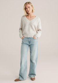 Maglione grigio chiaro a maglia con scollo a V e un motivo circolare texturizzato, abbinato a jeans a gamba larga blu chiaro. Scarpe marroni.