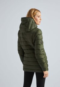 Veste matelassée vert foncé avec capuche, comportant des sections piquées, des poches latérales et un patch logo sur la manche gauche.