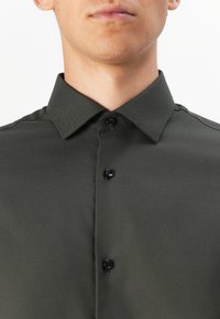 Mørk grønn skjorte med spredt krage, med svarte knapper og en glatt tekstur. Enkelt design uten synlige mønstre.
