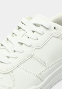 Λευκό δερμάτινο αθλητικό παπούτσι με στρογγυλή μύτη, υφή στις πλευρές και επίπεδα βαμβακερά κορδόνια; μινιμαλιστικός σχεδιασμός χωρίς ορατά λογότυπα.