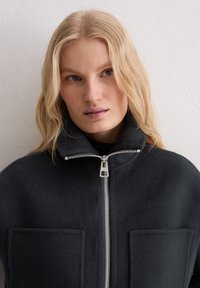 Ciemna wełniana kurtka z wysokim kołnierzem, przednim suwakiem i dwoma dużymi kieszeniami naszywanymi. Gładka faktura i dopasowany krój.