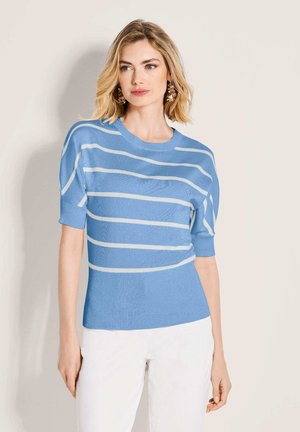 Frau mit schulterlangen blonden Haaren, die ein blau-weiß gestreiftes kurzärmeliges Oberteil und weiße Hose trägt und vor einem schlichten Hintergrund steht.