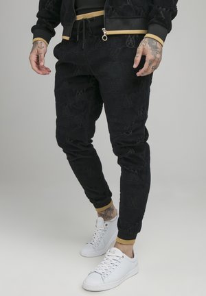 Homme portant un jogger noir à motifs avec un liseré doré et des baskets blanches, montrant des mains et chevilles tatouées sur un fond uni.