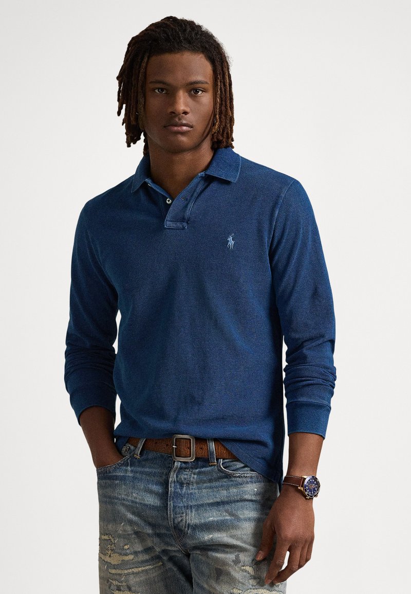 Μπλε σκούρο πουκάμισο πόλο με μακριά μανίκια, υφή υφάσματος, γιακάς με δύο κουμπιά και κεντημένο λογότυπο στο στήθος, σε συνδυασμό με σκισμένα μπλε τζιν.