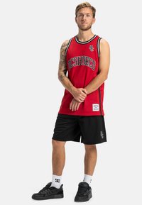 Červený sieťový basketbalový dres s čiernymi detailmi, s bielym a sivým textom loga. V páre s čiernymi sieťovanými šortkami a čiernymi teniskami.