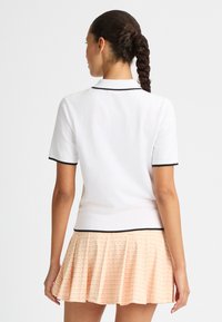 Kortärmad vit polo med svarta detaljer, ihop med en plisserad rosa kjol med geometriskt mönster. Slät tyg, figurnära skärning.