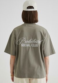 T-shirt oversized de culoare verde măsliniu, din material textil din bumbac, cu inscripție albă brodată "Prohibited Social Club" pe spate. Mâneci scurte, croială confortabilă.
