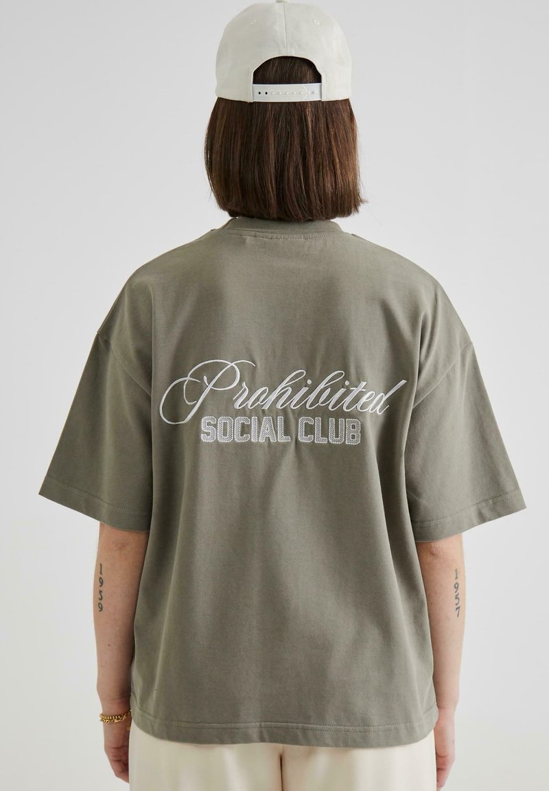 Olivenfarget oversized t-skjorte, laget av bomull, med hvit brodert tekst "Prohibited Social Club" på baksiden. Korte ermer, avslappet passform.