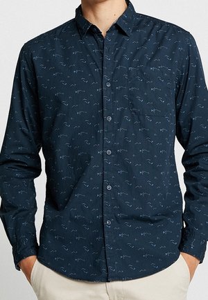 Marineblaue Hemdbluse mit dezentem hellblauem abstraktem Muster, Brusttasche, langen Ärmeln, getragen mit Händen in den Taschen einer beigen Hose.