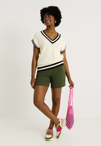 Top de punto blanco y negro a rayas con escote en V, pantalones cortos verdes y zapatillas multicolores. Sosteniendo una bolsa de malla rosa; atuendo casual de verano.