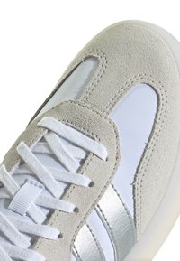 Sneaker en suède gris clair avec des lacets blancs, un panneau latéral synthétique blanc brillant et des rayures métalliques argentées. Semelle en caoutchouc texturée.