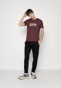Maroon t-shirt with prominent "BOSS" logo, paired with black tapered pants and multi-colored sneakers. Simple design, crew neckline, short sleeves.