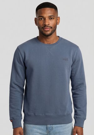 Uomo con capelli corti e barba che indossa una felpa blu a girocollo e jeans azzurri, in piedi davanti a uno sfondo grigio uniforme.