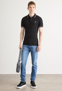 Polo noir avec des accents blancs, jeans bleus et baskets noires. Sac bandoulière gris. Le modèle se tient devant un fond neutre.