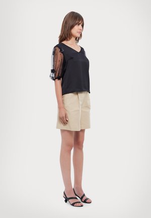 Schwarzes Satin-Oberteil mit transparenten schwarzen Puffärmeln mit floralen Akzenten, kombiniert mit beigen Shorts und schwarzen Riemchen-Absätzen. Glatte Textur.