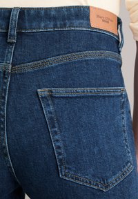 Jeans en denim bleu foncé avec une étiquette en cuir portant la mention "Marc O'Polo". Présente des coutures visibles et un design classique à cinq poches.