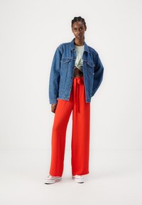 Jeansjakke med mønsterdesign, grønn knyttet topp og vide, lyse røde bukser. Hvite sneakers fullfører antrekket.