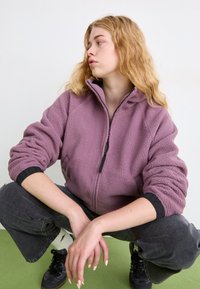 Chaqueta de forro polar con cremallera en morado claro y detalles en negro, con un cuello alto y puños elásticos. Combinada con pantalones gris oscuro y zapatillas negras.