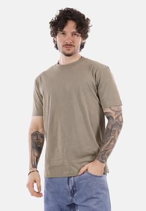 Uomo con capelli ricci che indossa una maglietta beige e jeans blu, mostrando tatuaggi dettagliati su entrambi gli avambracci, in piedi contro uno sfondo bianco.