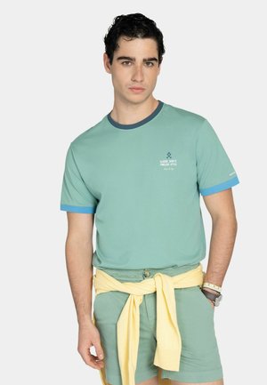 Joven con cabello rizado oscuro, vistiendo una camiseta verde, pantalones cortos a juego, un suéter amarillo atado a la cintura y un reloj de pulsera.