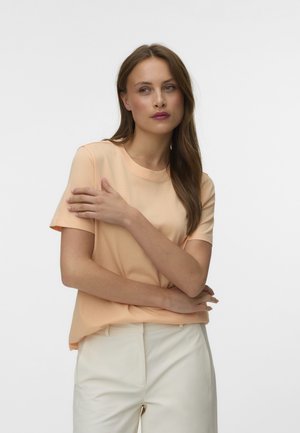 Femme aux longs cheveux bruns portant un t-shirt pêche à manches courtes et un pantalon blanc, les bras croisés, regardant sur le côté sur fond blanc.