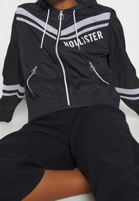 Mustahoodattu tuulenpitävä takki, jossa on valkoiset raidat, vetoketjulliset taskut ja rintamuksessa brändi "HOLLISTER" tulostettuna. Sileä kankaan pinta.