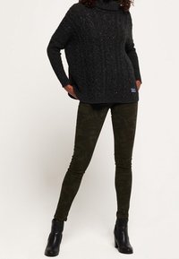 Persoon in een donkergrijze kabelgebreide coltrui, met camouflagemotief skinny pants en zwarte hielen enkelboots, staand met handen in de zakken.