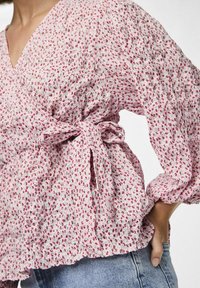 Blusa floreale rosa con delicati motivi rossi e verdi, texture smock, scollatura a V profonda e dettaglio di vita legata. Maniche lunghe e puffate.
