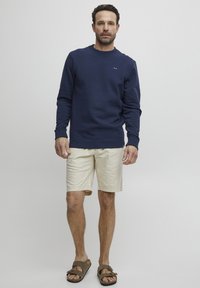 Felpa blu navy con collo rotondo, maniche lunghe e dettaglio del logo; abbinata a pantaloni corti crema e sandali marroni. Tessuto morbido e texturizzato.